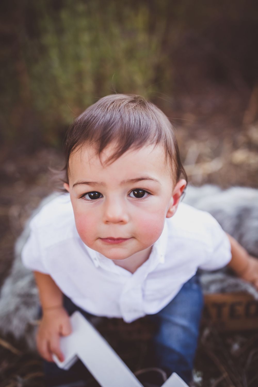 Small child in white shirt and blue pants sitting and looking up at camera with a sweet expression outdoors.