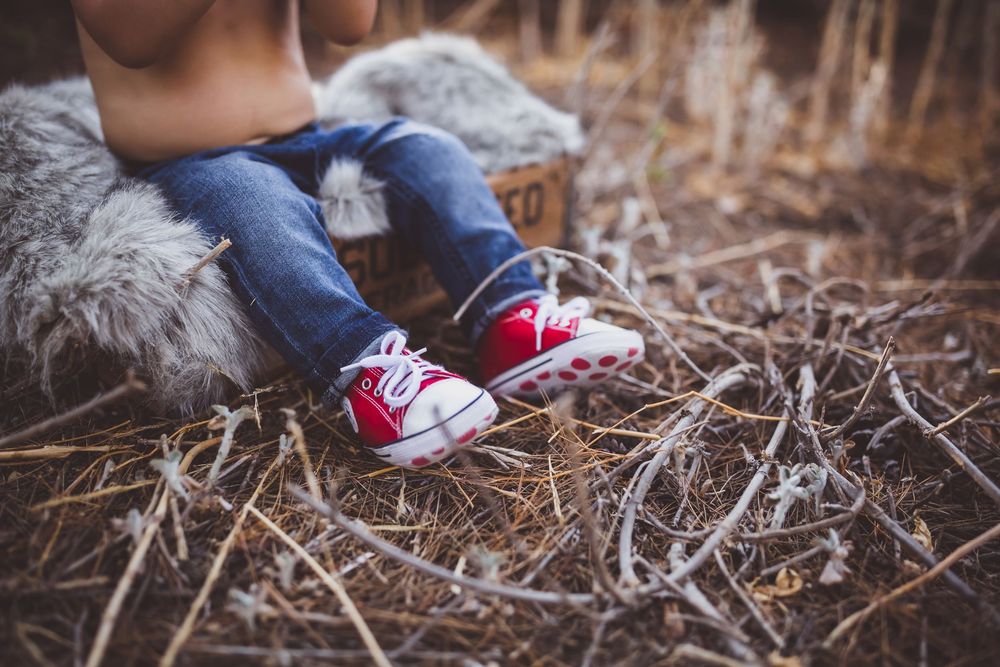 Small feet wearing red and white sneakers sit on dried twigs and grass in an outdoor autumn setting.