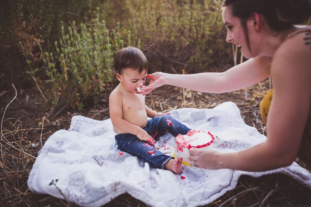 Infant in white dress lying on ground being photographed during outdoor photo session.