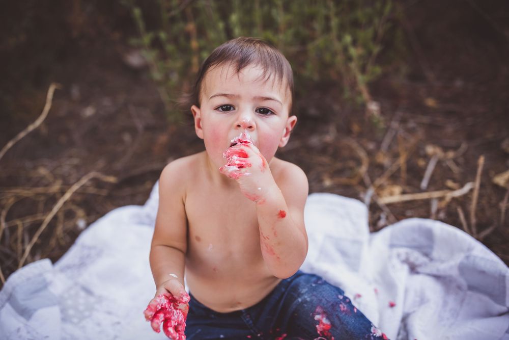 Child sits in tulle with messy red frosting around mouth during cake smash photo shoot.