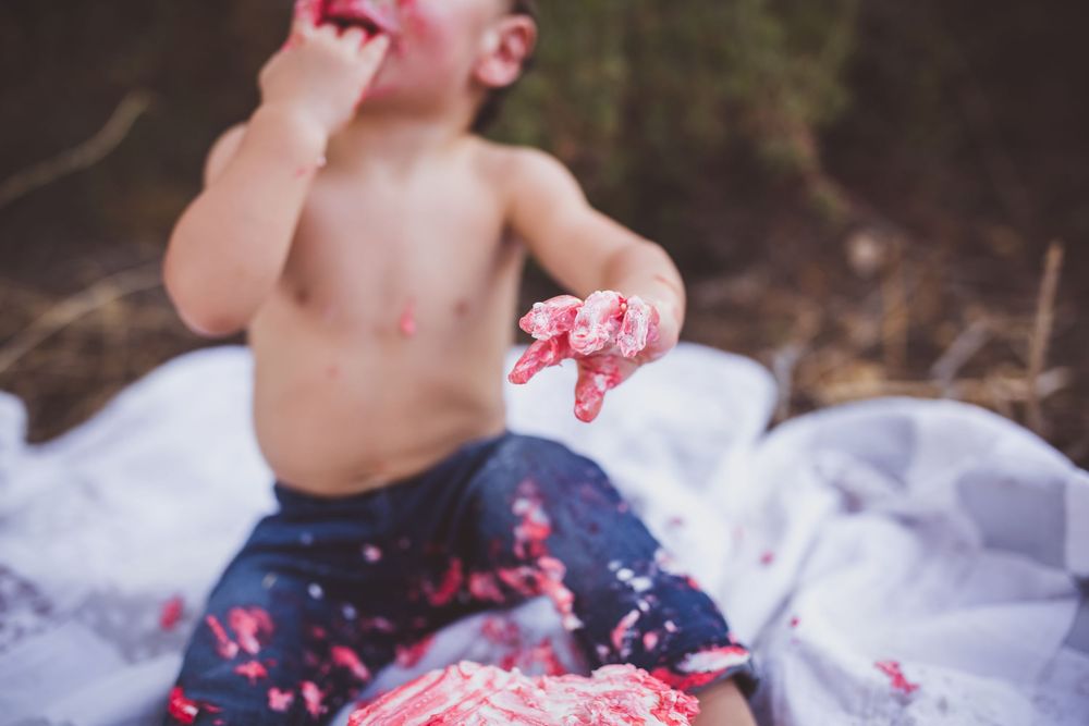 Baby in denim shorts sits on blanket outdoors messily eating cake during first birthday smash cake session.