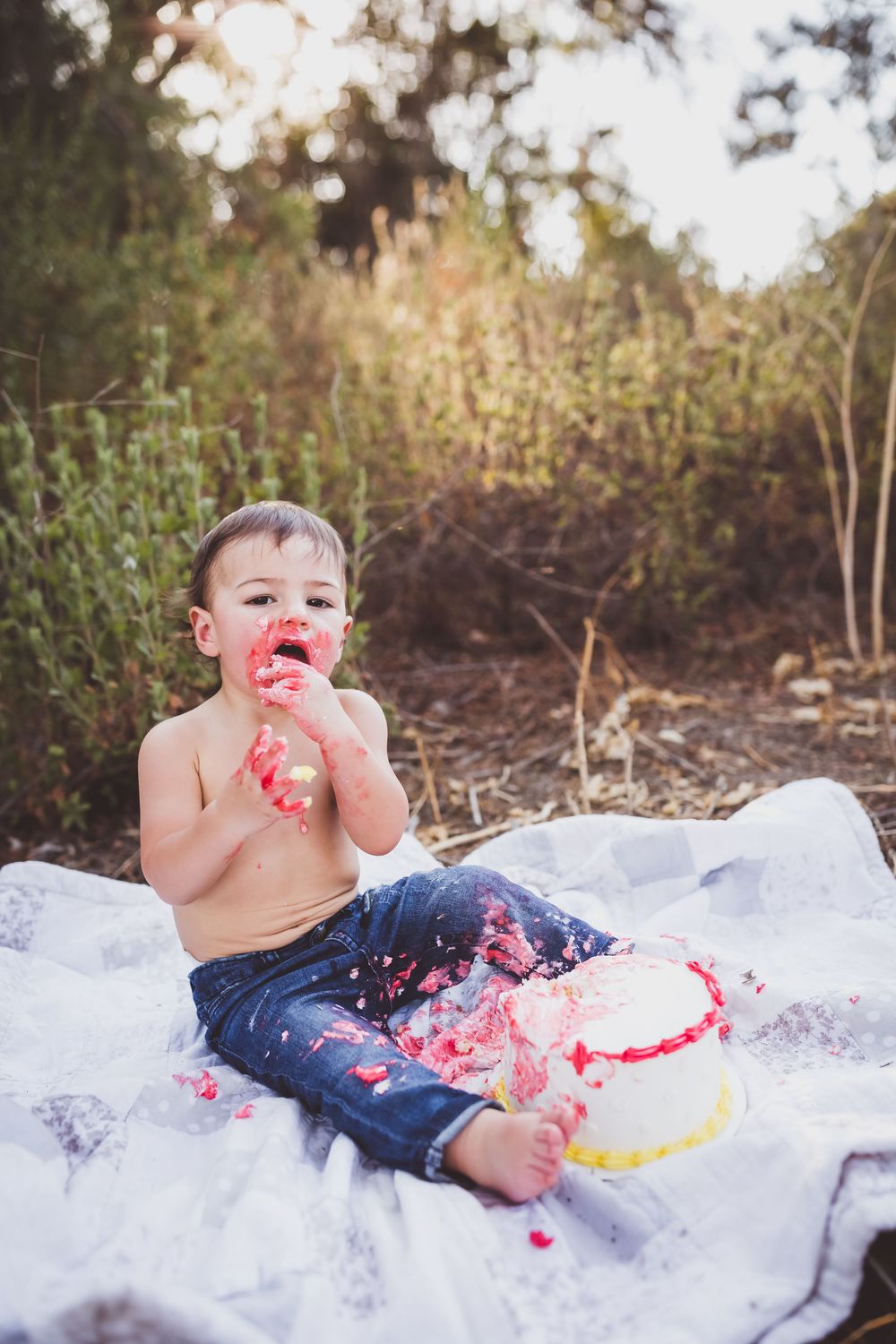 Baby in jeans smashing cake and frosting during first birthday cake smash photo session on white blanket outdoors.