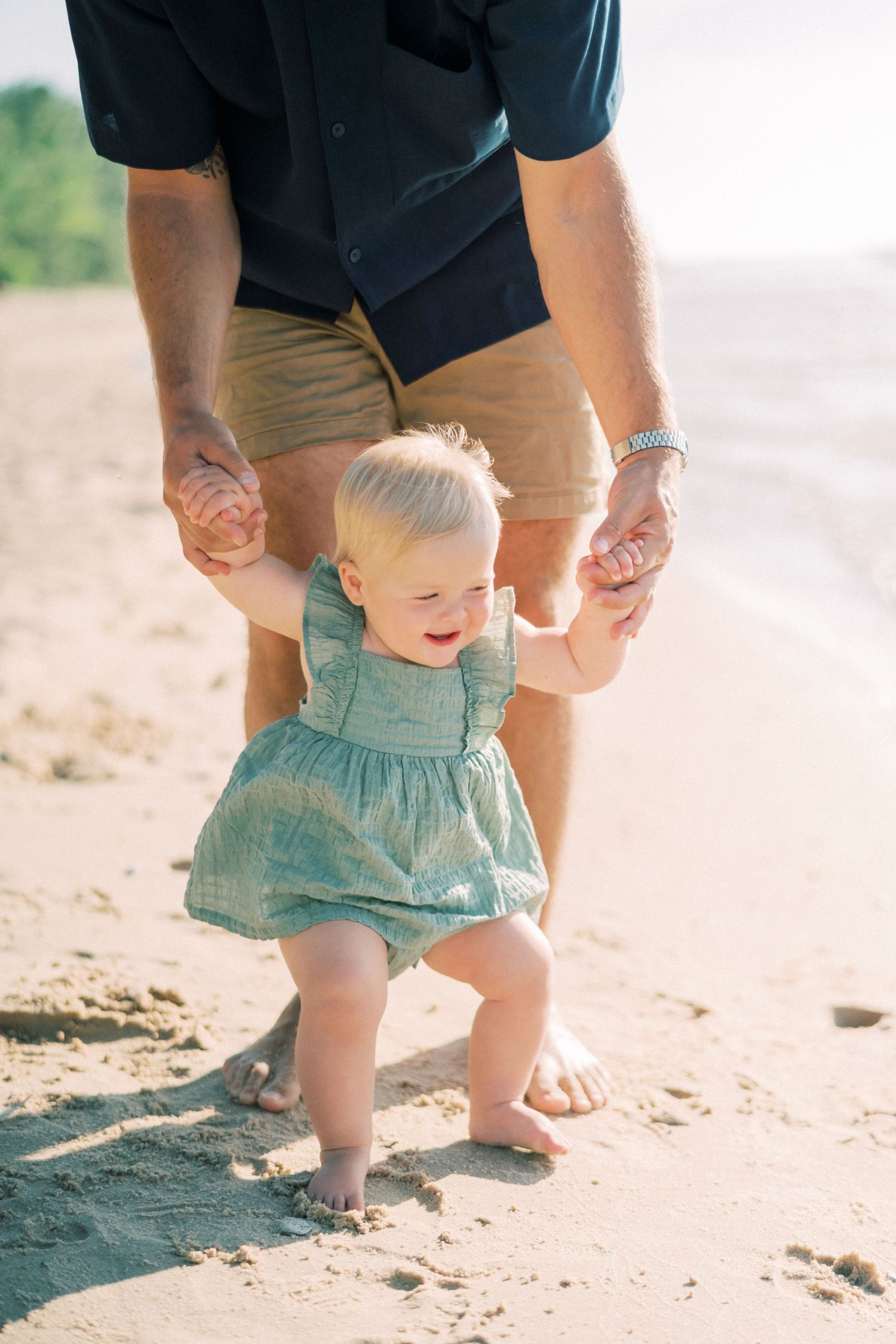 Small child in turquoise dress takes first steps on beach while holding adult's hands for support on sunny day.