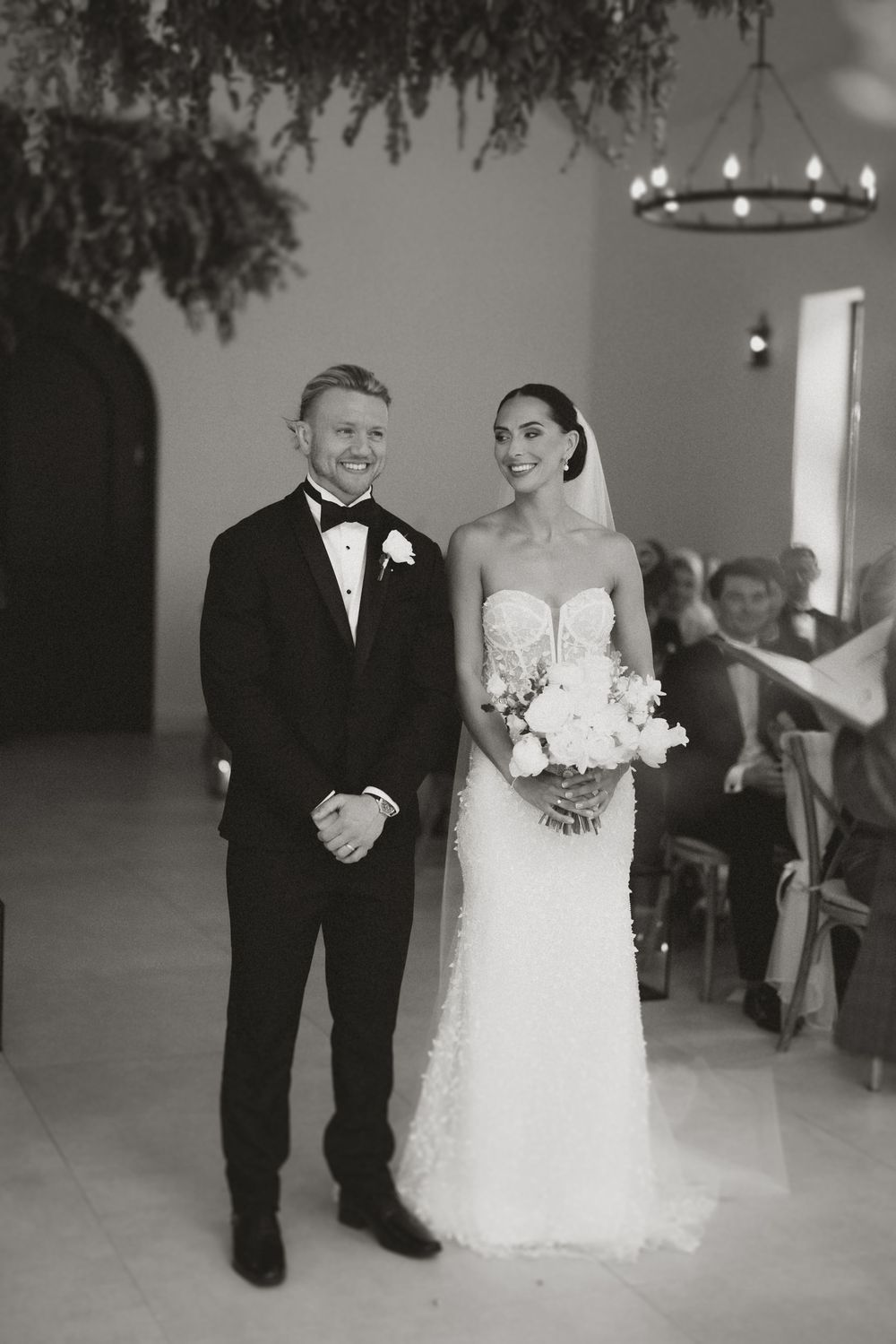 A black and white wedding photo showing a couple at their ceremony, the bride in a strapless gown and groom in tuxedo.