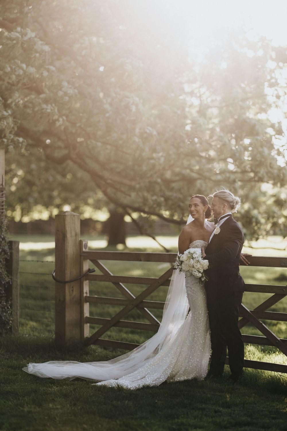 Wedding couple stands together by a rustic wooden gate in a sunlit garden during golden hour.