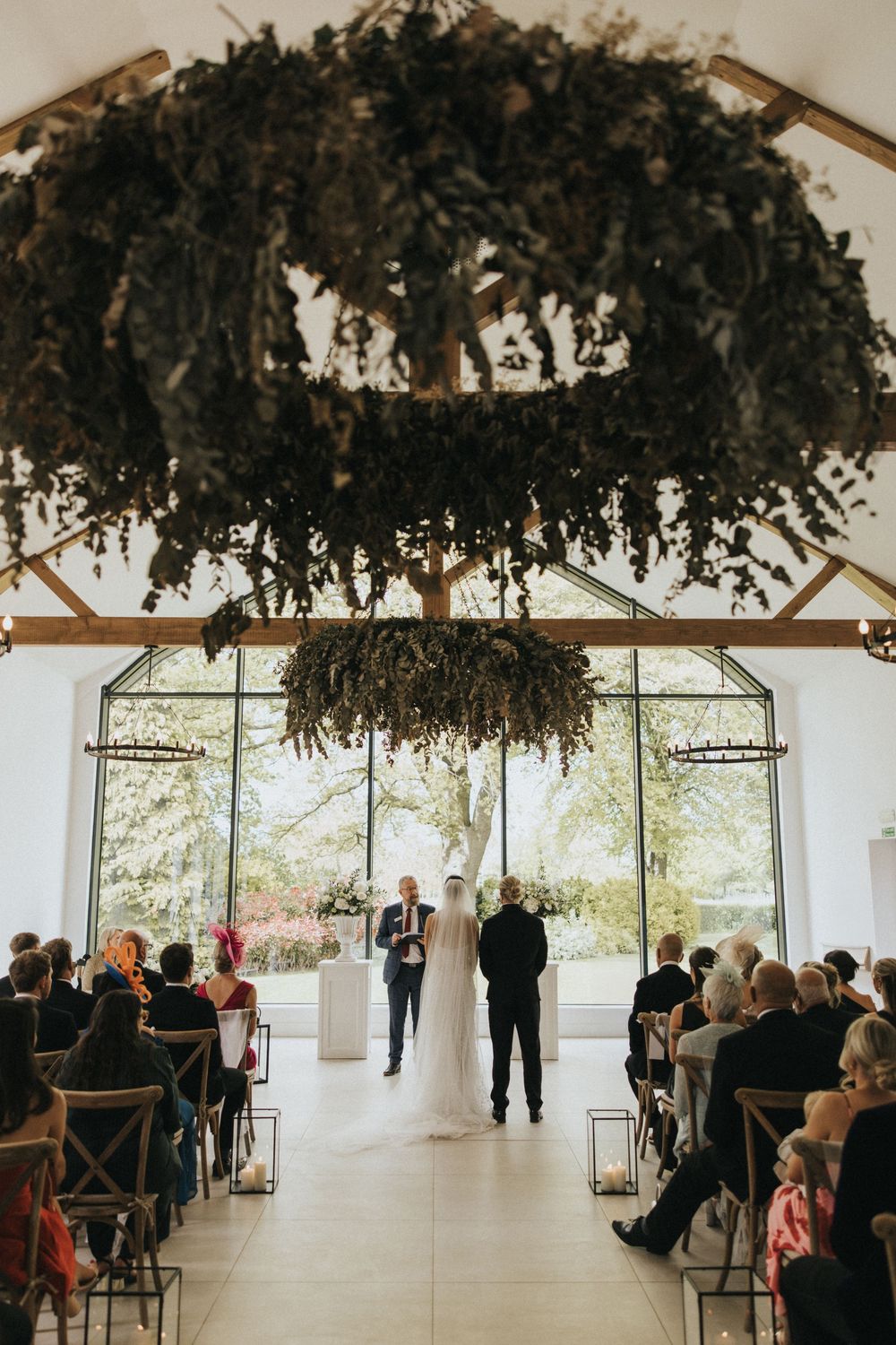 Indoor wedding ceremony setup with dried floral ceiling decorations and rows of wooden chairs.
