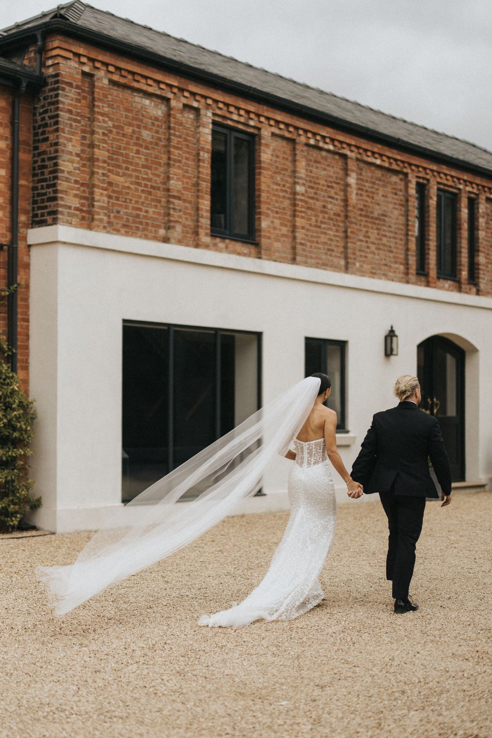 Wedding couple walks outside a rustic brick venue as bride's long veil flows in the breeze.