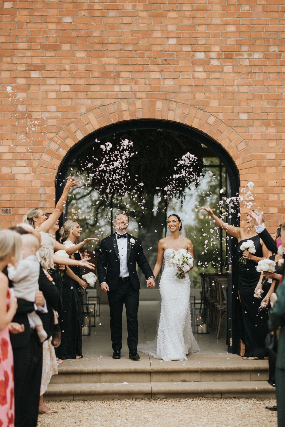 Wedding couple stands on church steps while guests toss confetti in celebration under a brick archway.