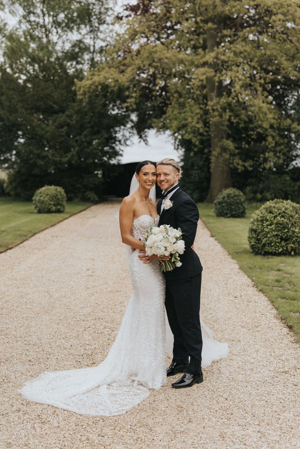 A couple poses on a gravel path surrounded by manicured grounds, the bride wearing a fitted lace gown holding white flowers.