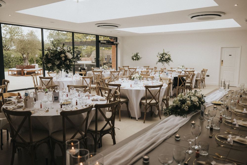 Light-filled reception venue with round tables, cross-back chairs, and elegant floral centerpieces in a modern white space.