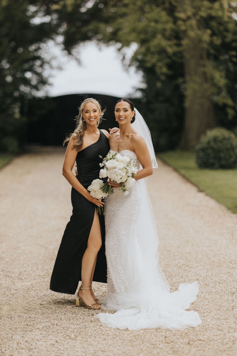 A bride in a white wedding gown laughs with someone in black attire on a gravel path outdoors.