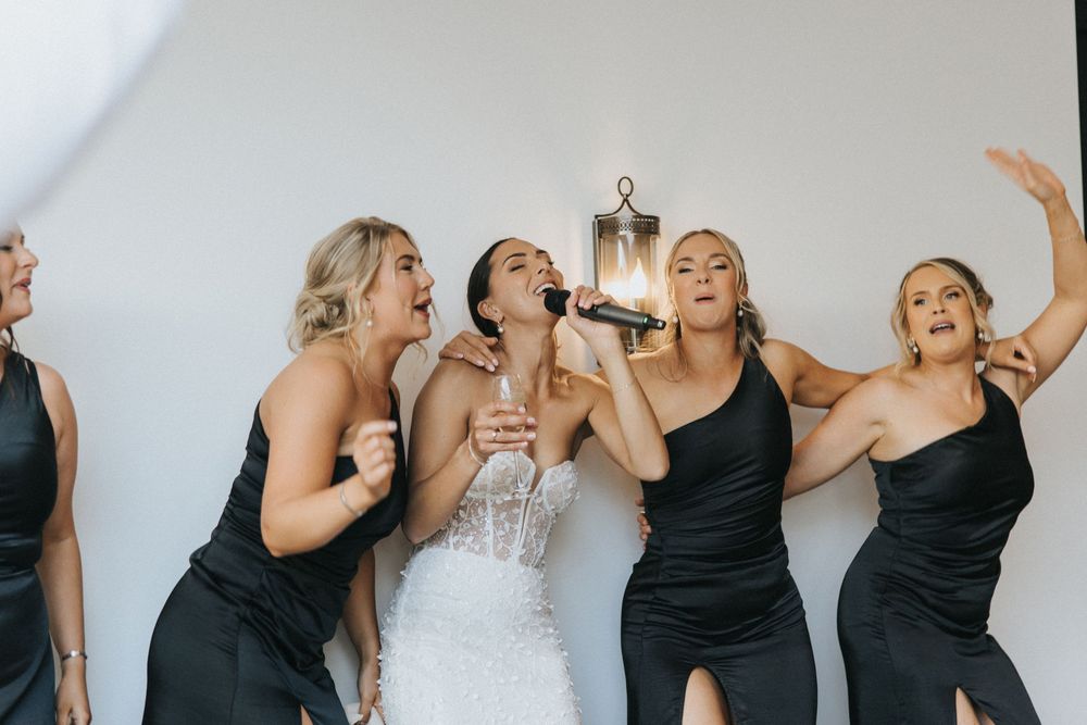 A bride and bridesmaids in black dresses laugh joyfully together during a candid wedding photo moment.