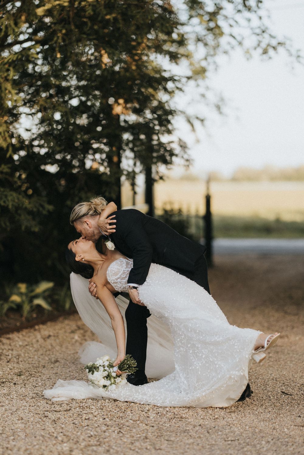 Romantic couple share a dip kiss on their wedding day with a dreamy sunset background and flowing white dress.