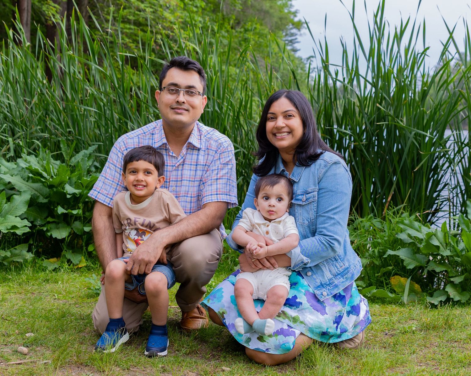 A family of four poses together outdoors in front of tall green grass on a sunny day.