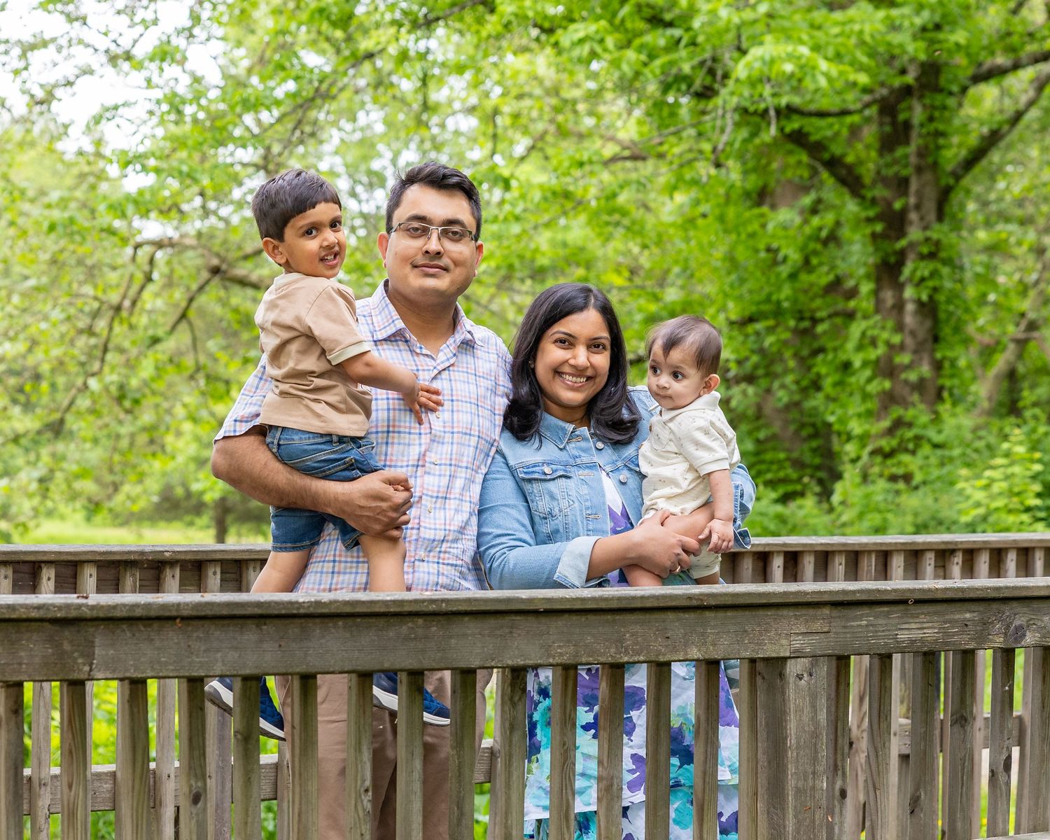 A family shares joyful moments together while posing on a wooden fence in a lush green outdoor setting.