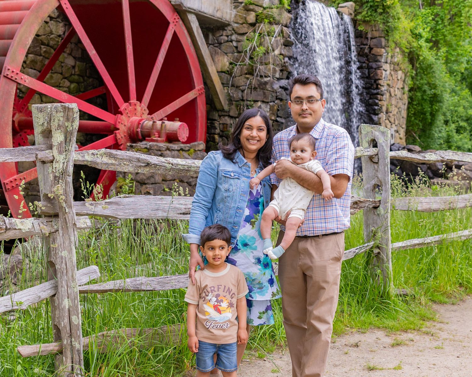 Family poses near red water wheel and waterfall at rustic mill with wooden fence in rural outdoor setting.