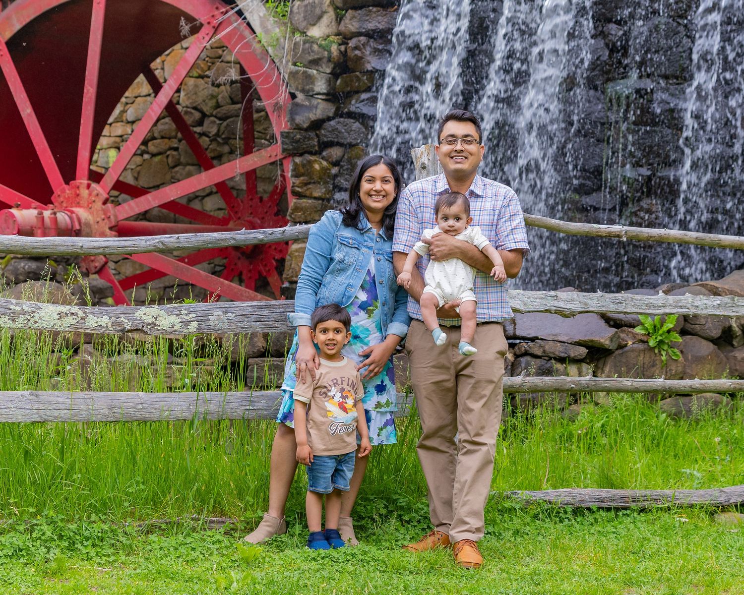A family stands together by a rustic water wheel and waterfall at a historic mill site surrounded by green grass.