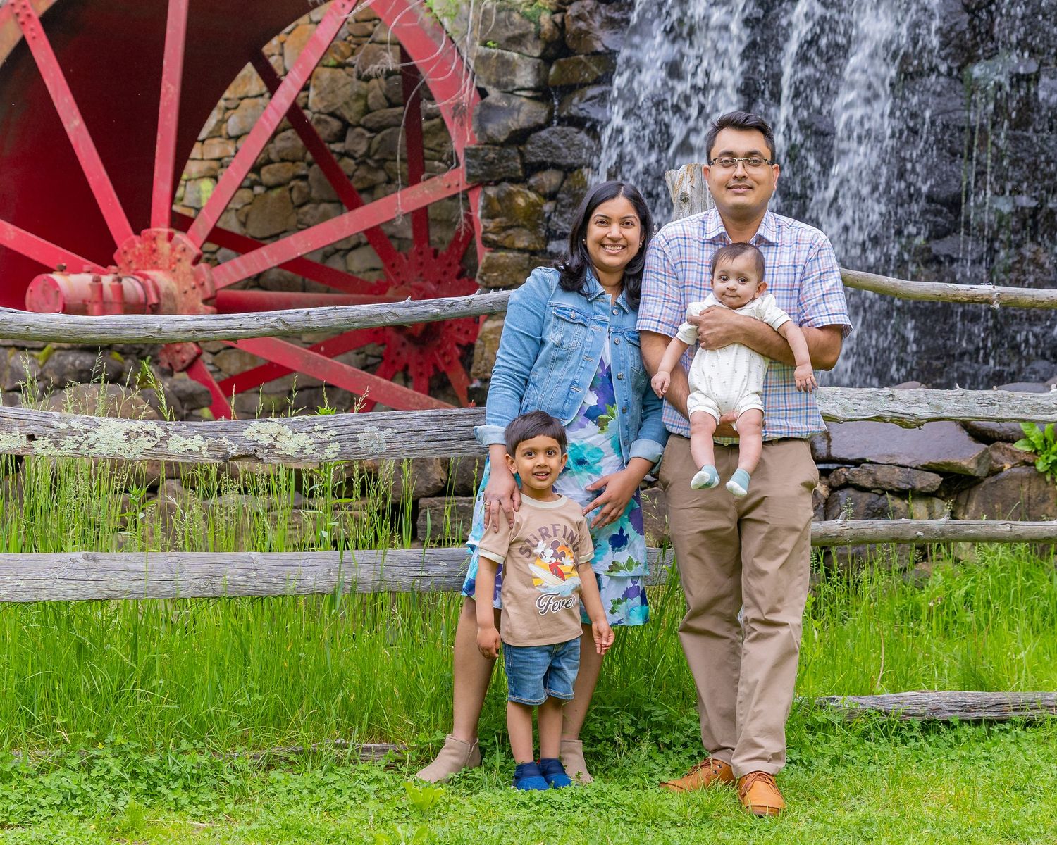 A group poses together against a cascading waterfall backdrop near a red waterwheel in a scenic garden setting.