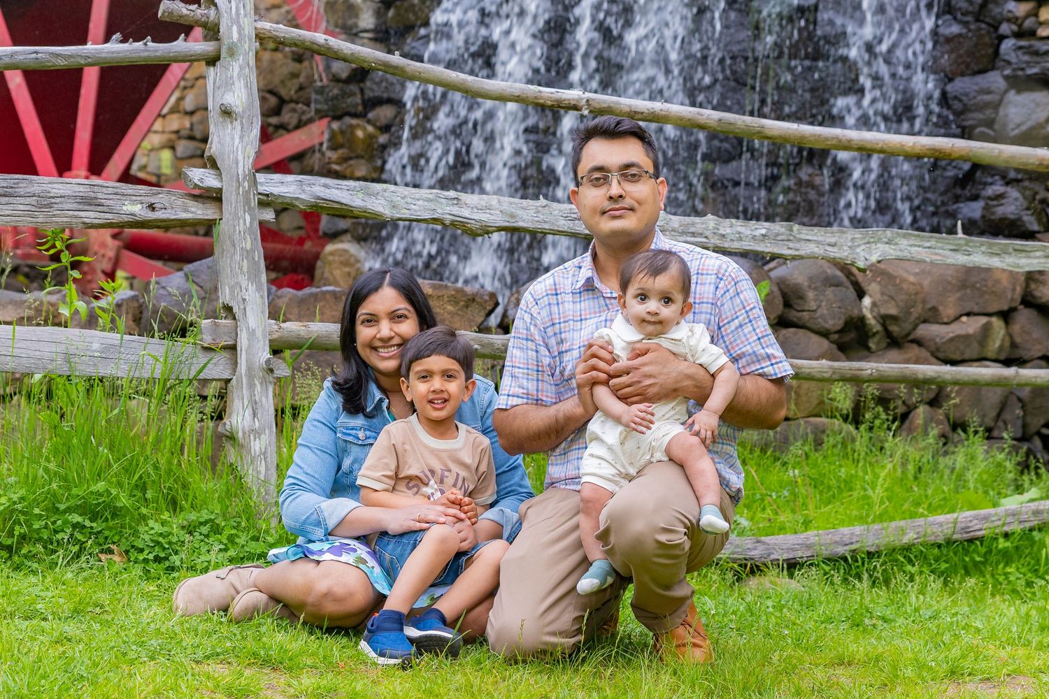 A family sits together on grass near a wooden fence and waterfall on a sunny day in nature.