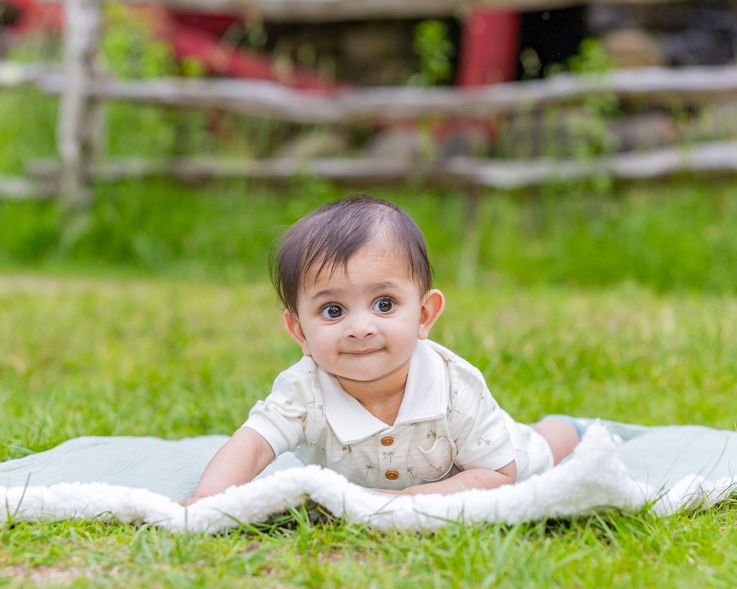 Baby wearing white clothing lies on a blanket outdoors in grassy setting during playtime.