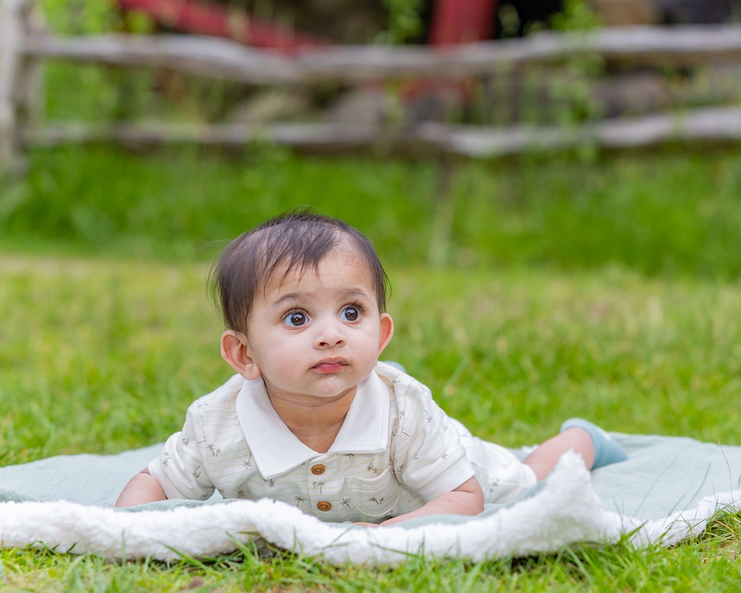 Baby in a white outfit lying on a blanket in green grass outdoors.