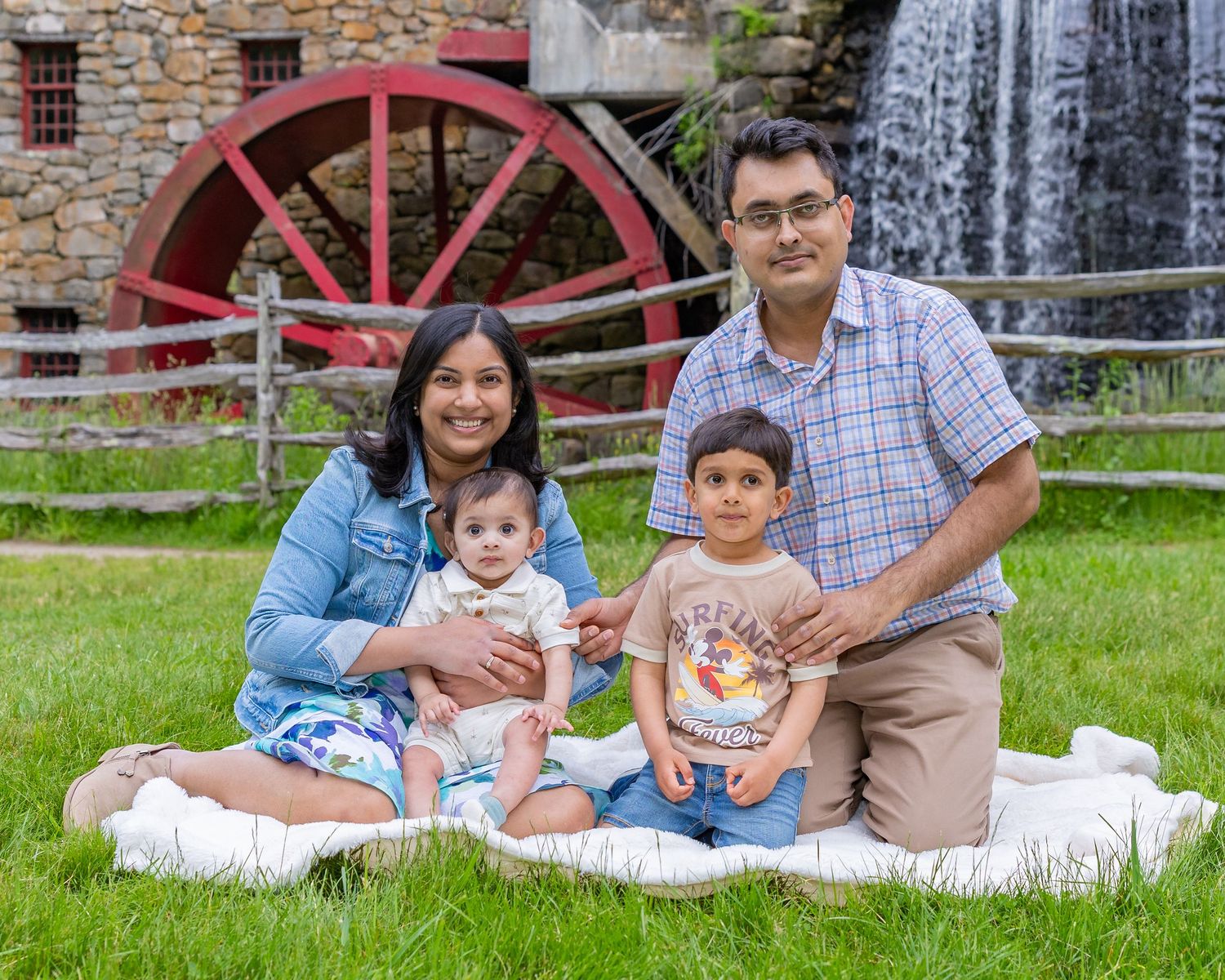 Family sitting together outdoors on grass near historic red water wheel and brick wall.