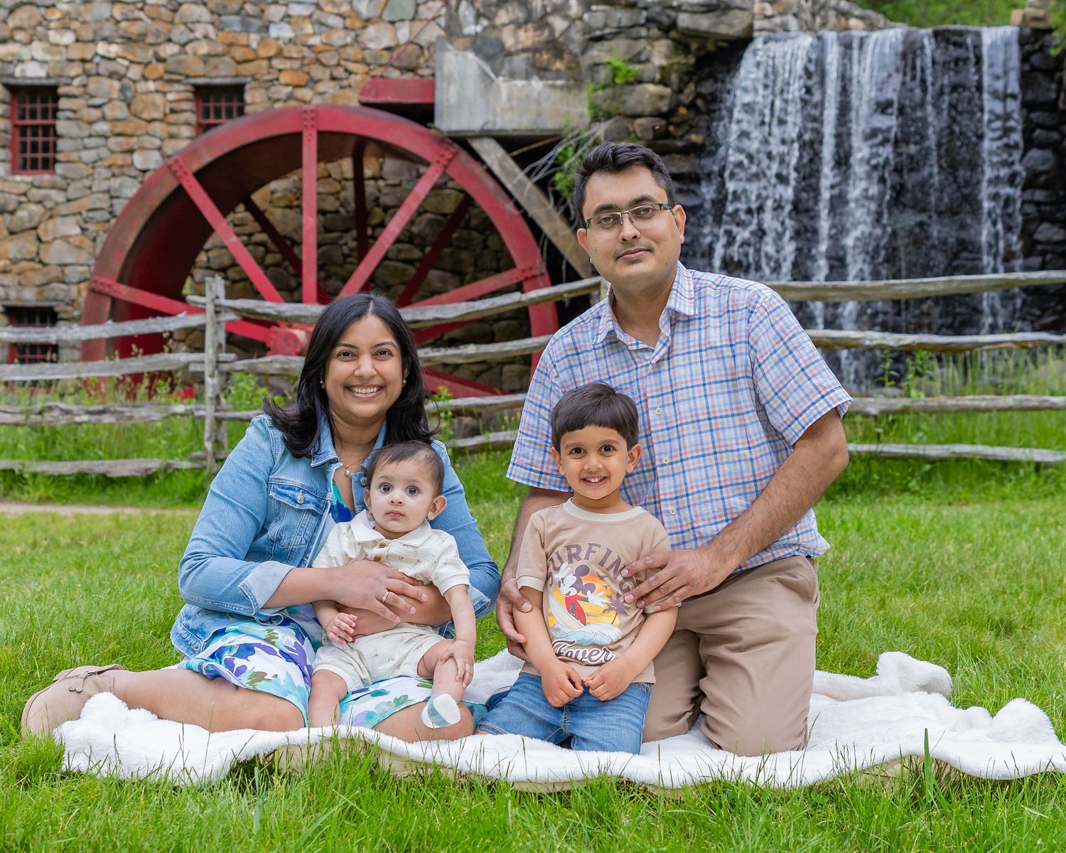 Family enjoys a relaxing summer day on the grass near an old stone mill with a red water wheel in the background.