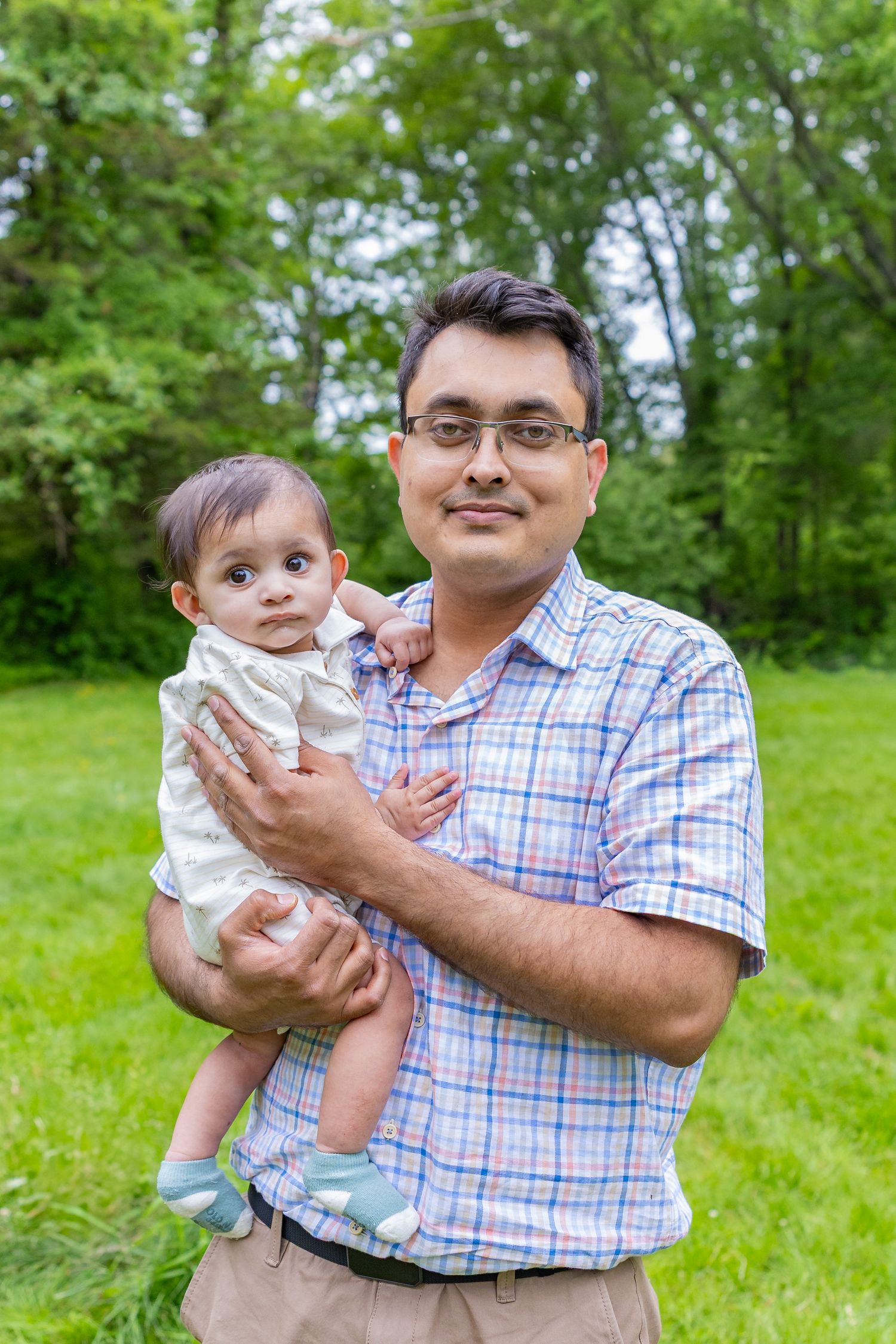A plaid-shirted person holds a baby in white clothing while standing in a lush green outdoor setting.