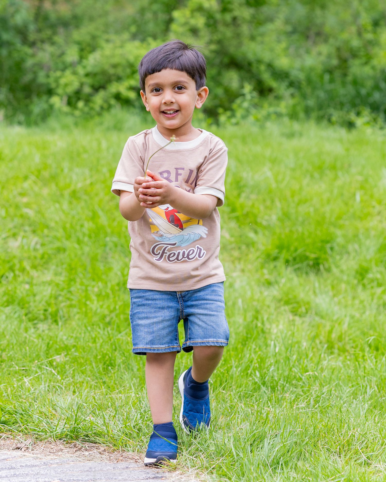 Small child wearing beige shirt and denim shorts runs playfully on grass path during outdoor summer activity.