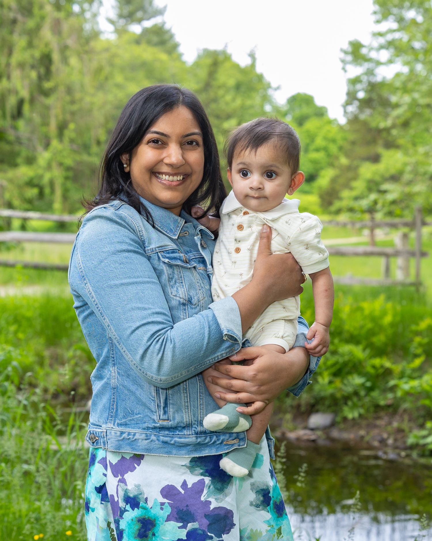 A parent and baby stand together in a green garden setting with a pond in the background on a sunny day.
