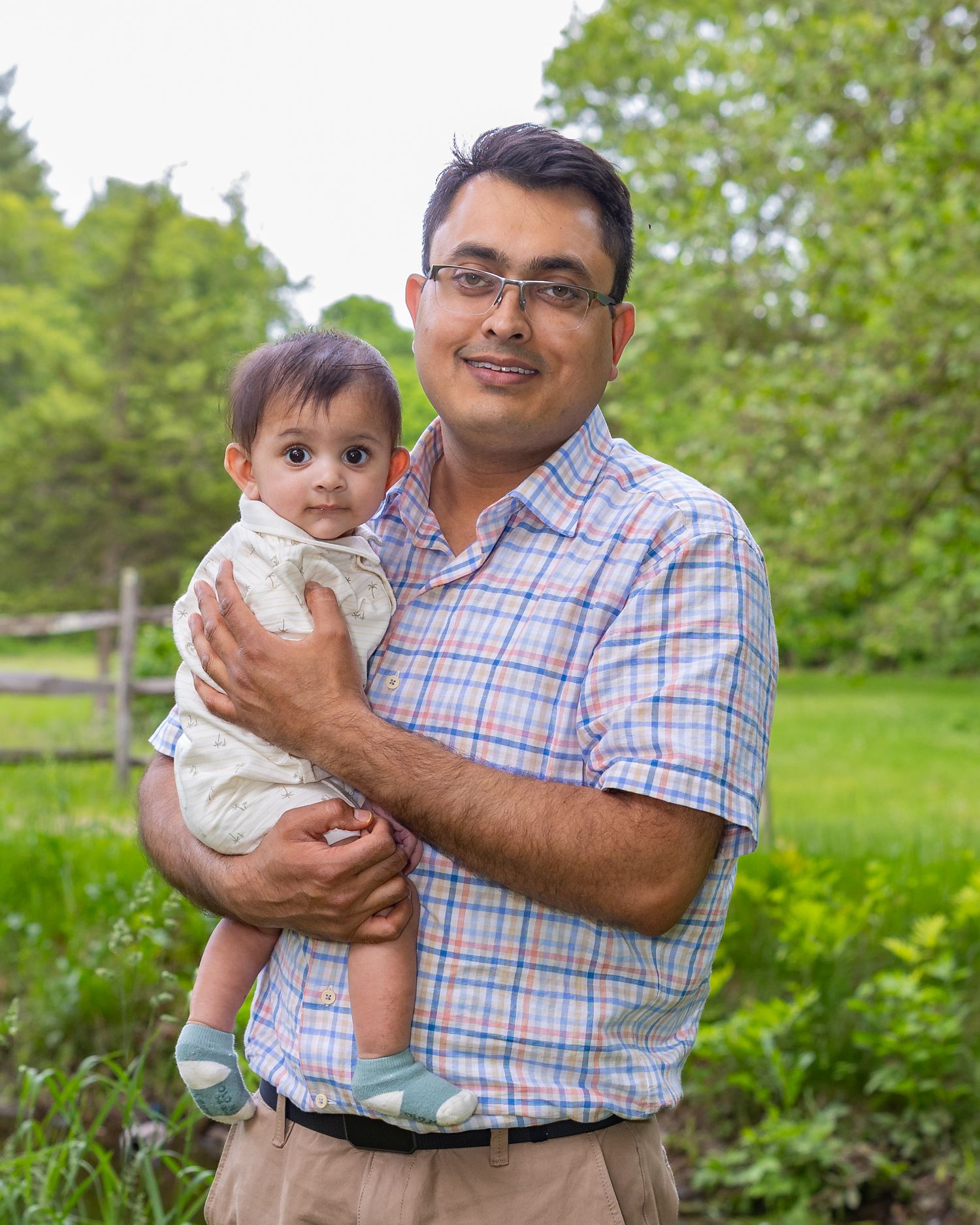 A parent and baby stand together outdoors in a garden area with green trees and grass in the background.