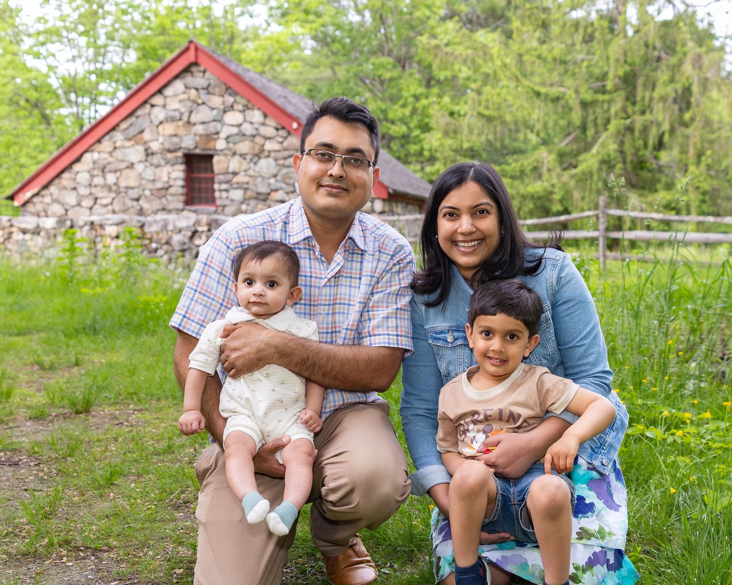 A family poses together outdoors in front of a rustic stone building surrounded by green spring foliage.