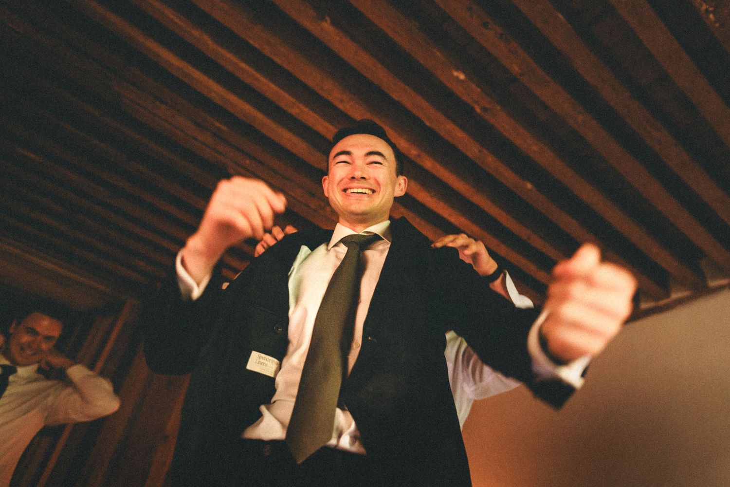 A person in formal attire dancing enthusiastically under wooden ceiling beams.