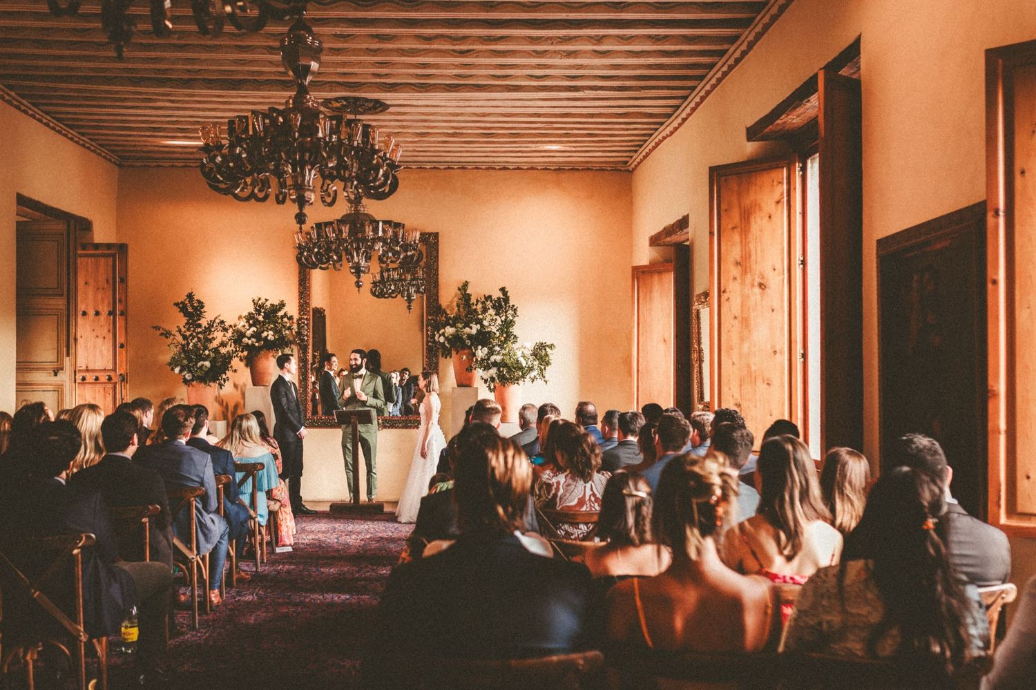 Guests seated in an elegant reception hall with wooden ceiling beams and chandeliers.
