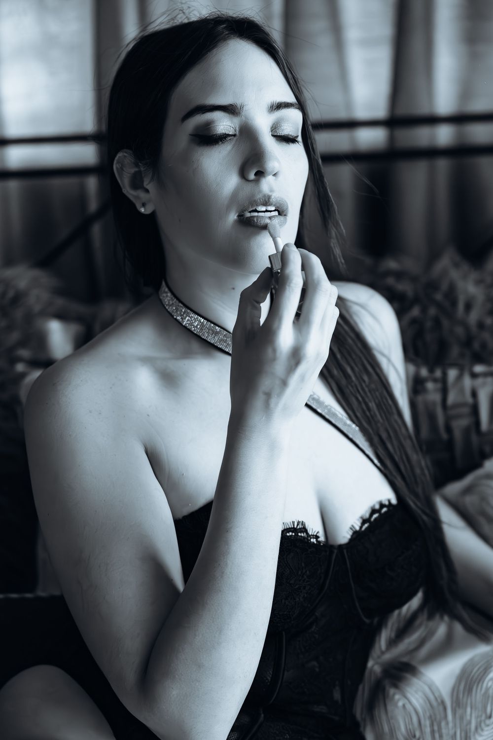 Elegant black and white portrait of a person wearing a corset and choker, applying lipstick with eyes closed.