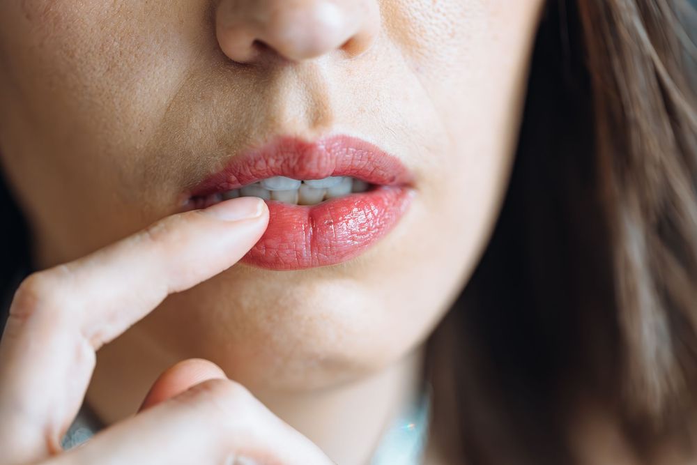 Close up of someone wearing red lipstick touching their finger to glossy lips against dark background.