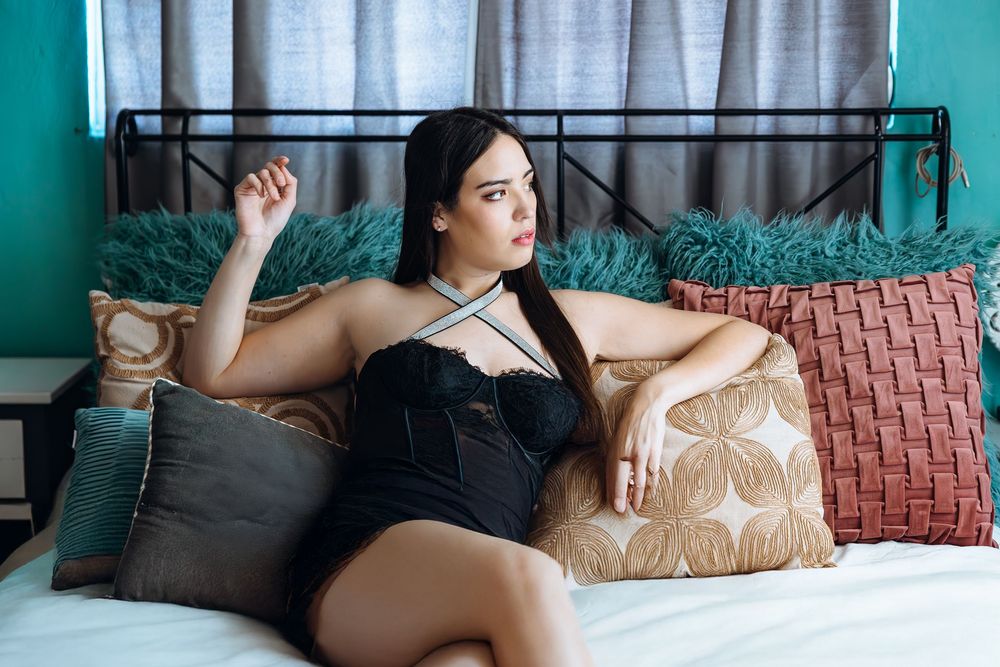A person relaxes on a bed with decorative pillows against a teal wall wearing black lingerie.