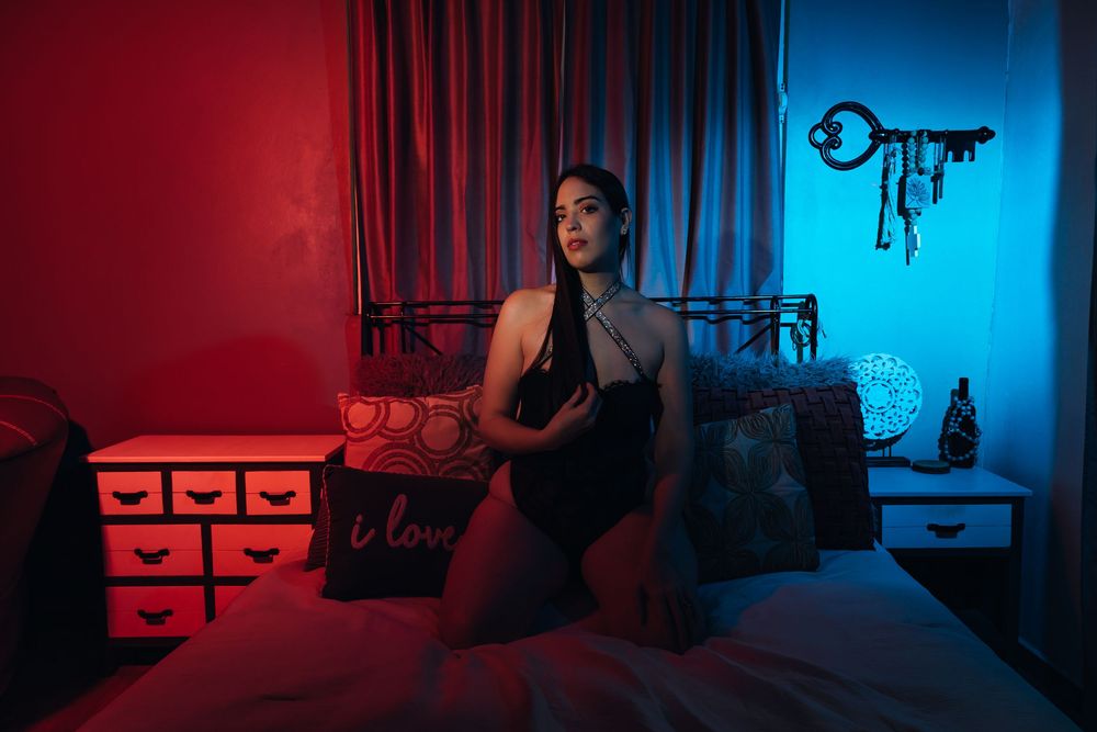 A bedroom lit with dramatic red and blue lighting features a figure seated on a bed near decorative storage drawers.