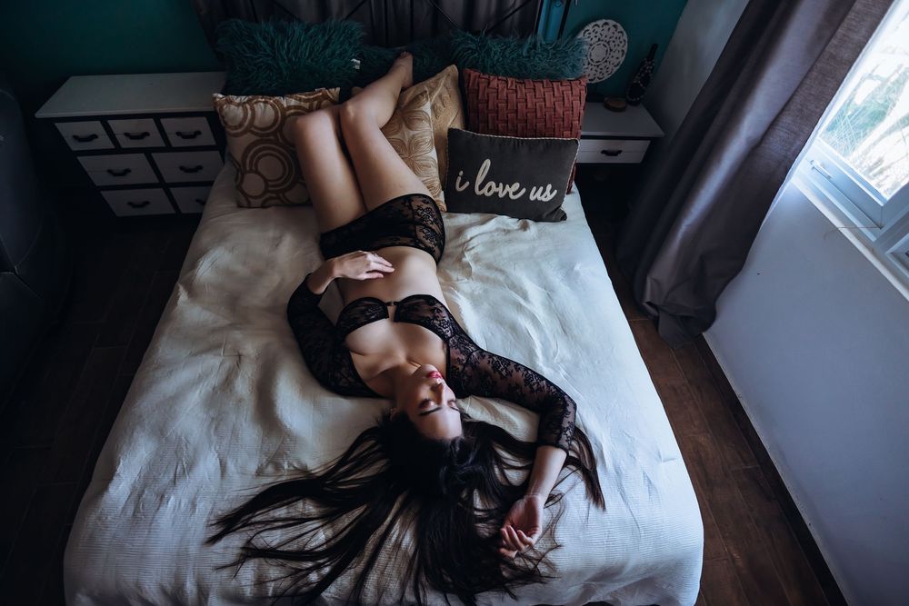 A person in black attire lying on white bedding with long dark hair spread out across the bed.