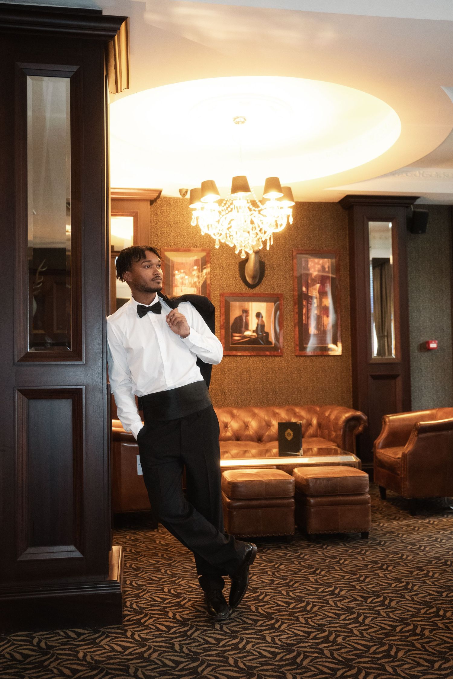 Elegant lounge interior with crystal chandelier, leather seating, and formal-dressed individual in bowtie against ornate decor.