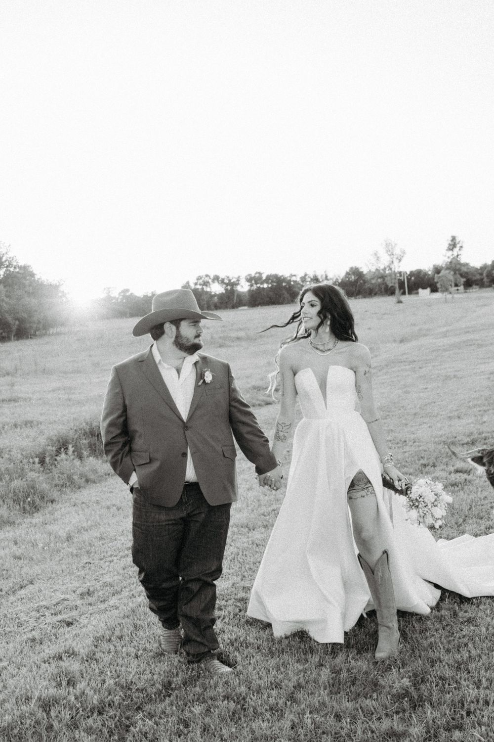 A couple in western wedding attire walk hand-in-hand through a sunlit field.