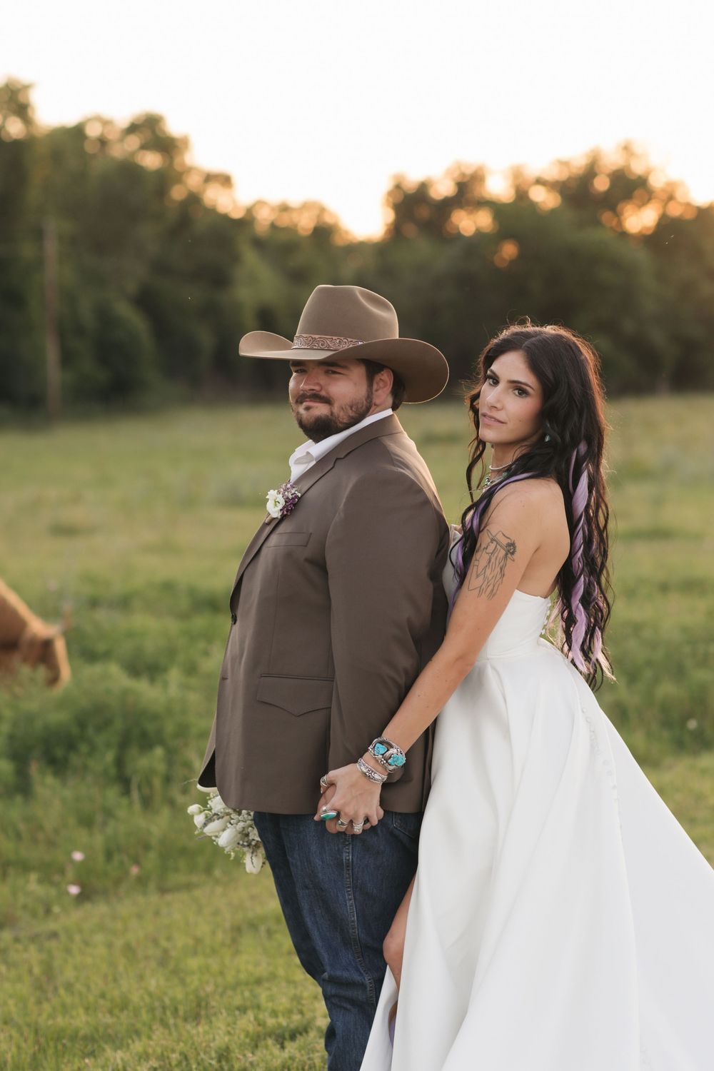A bride in a white dress and groom in brown suit pose together in a rustic outdoor setting at sunset.