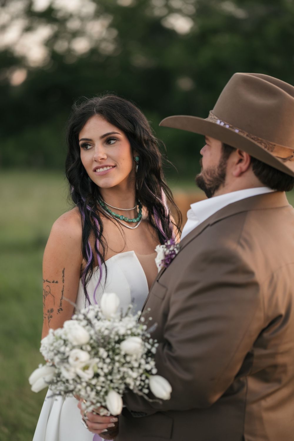 A romantic wedding photo featuring a couple in western attire with the bride holding a white floral bouquet outdoors.