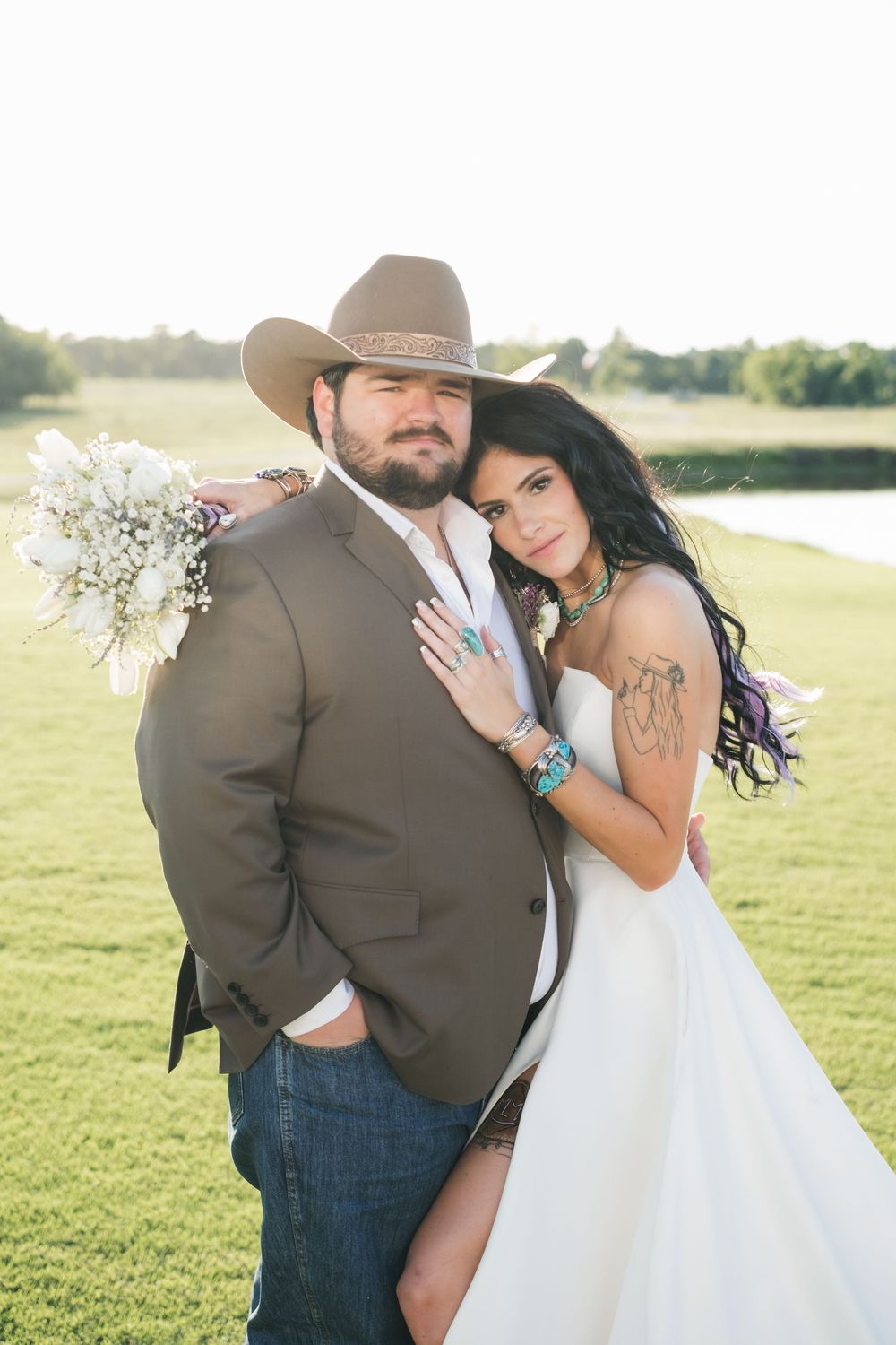 A country wedding couple poses together on a grassy field at sunset, with the bride holding a delicate bouquet.