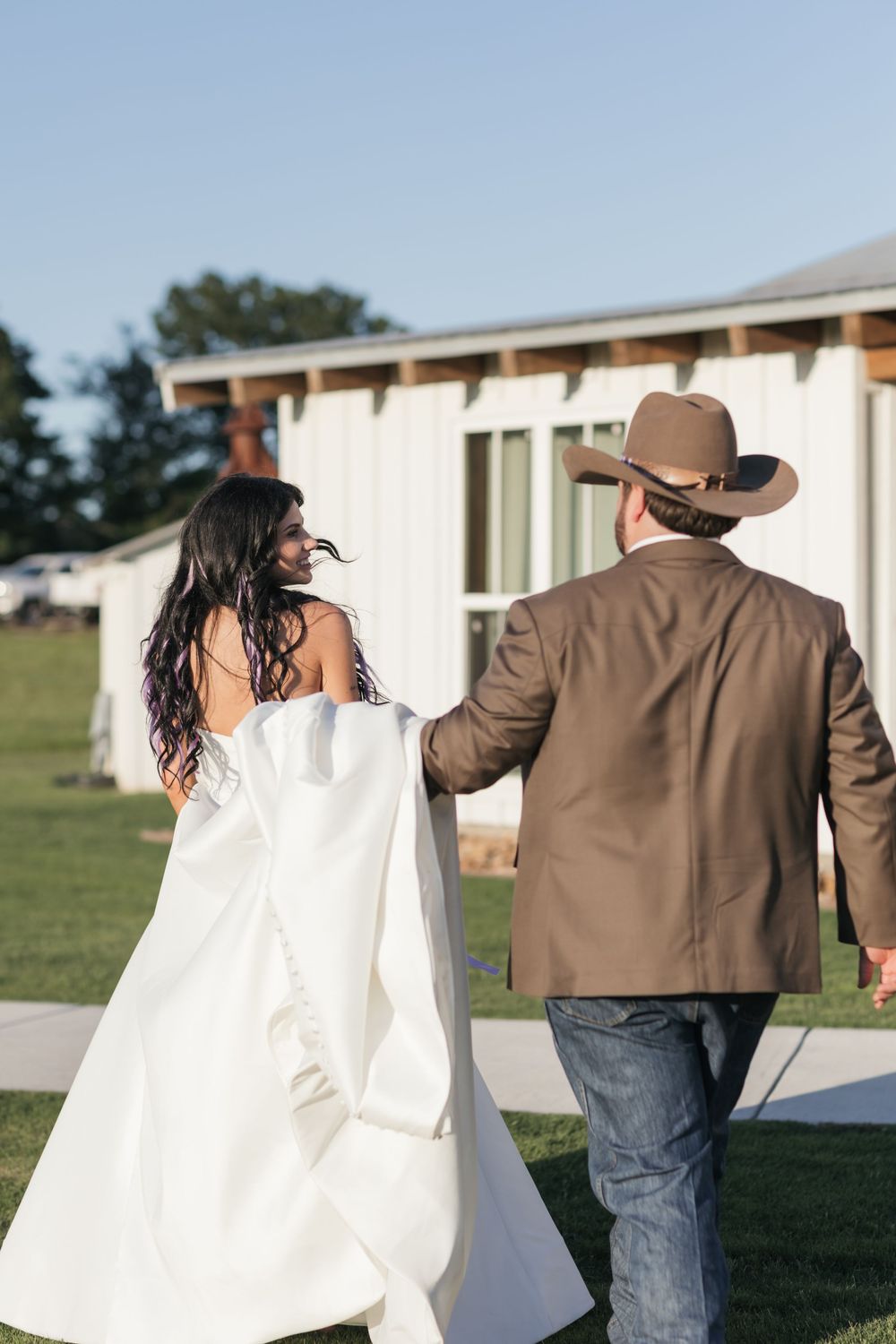 A couple walking together outdoors, with the bride in a flowing white dress and the groom wearing a brown jacket and cowboy hat.