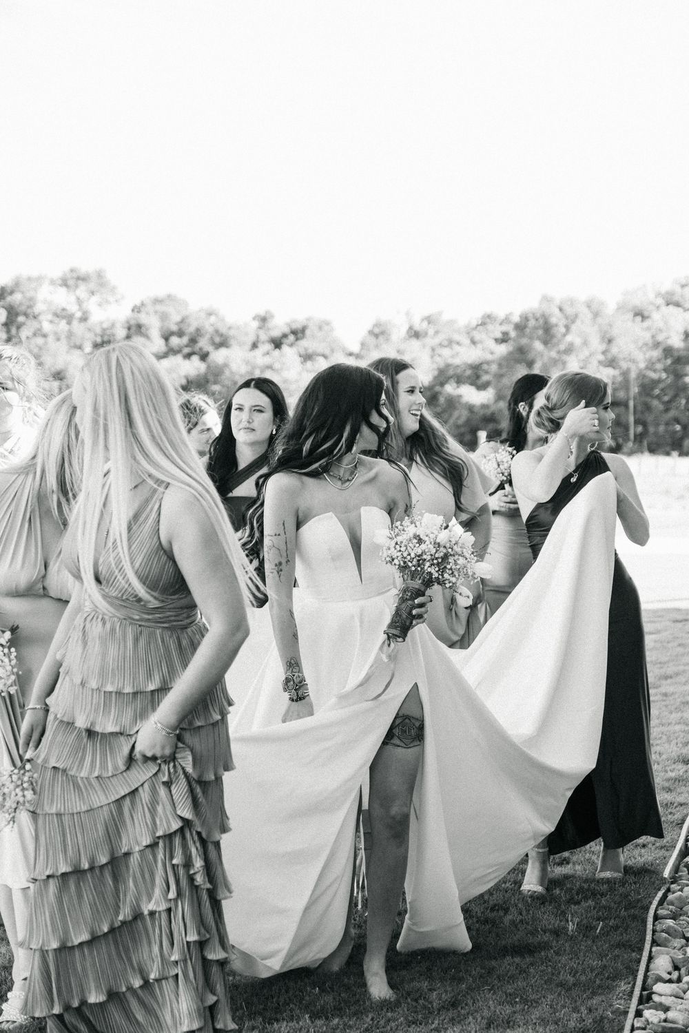 Group photo of bridesmaids in lavender dresses and a bride in white posing outdoors at a wedding.