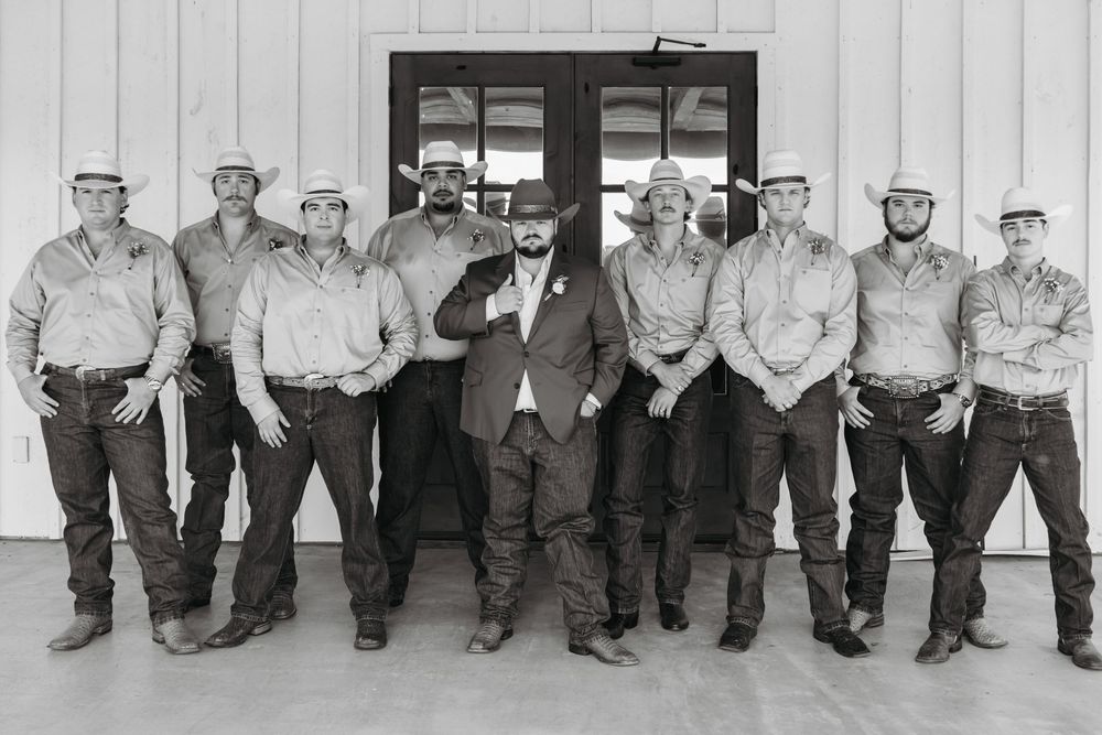 A group of people in casual western attire and cowboy hats line up against a light-colored wall for a black and white photo.