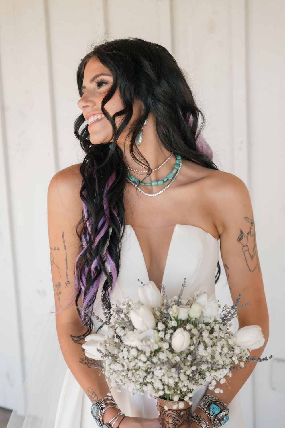 Bride with purple-streaked hair holds white bouquet while wearing turquoise jewelry and strapless wedding gown.