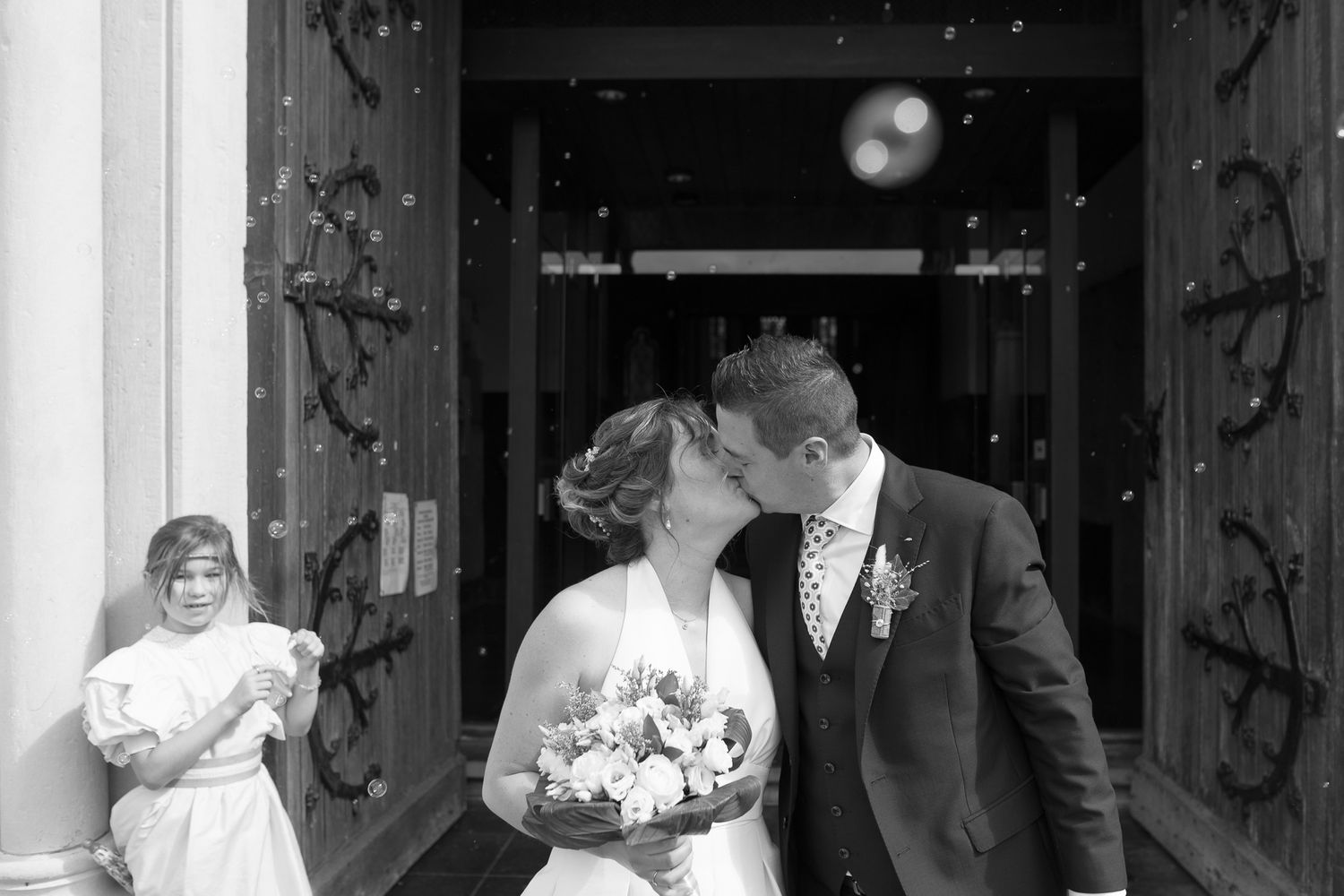 Black and white photograph capturing a romantic moment at a church entrance with bridal flowers and decorative vines.