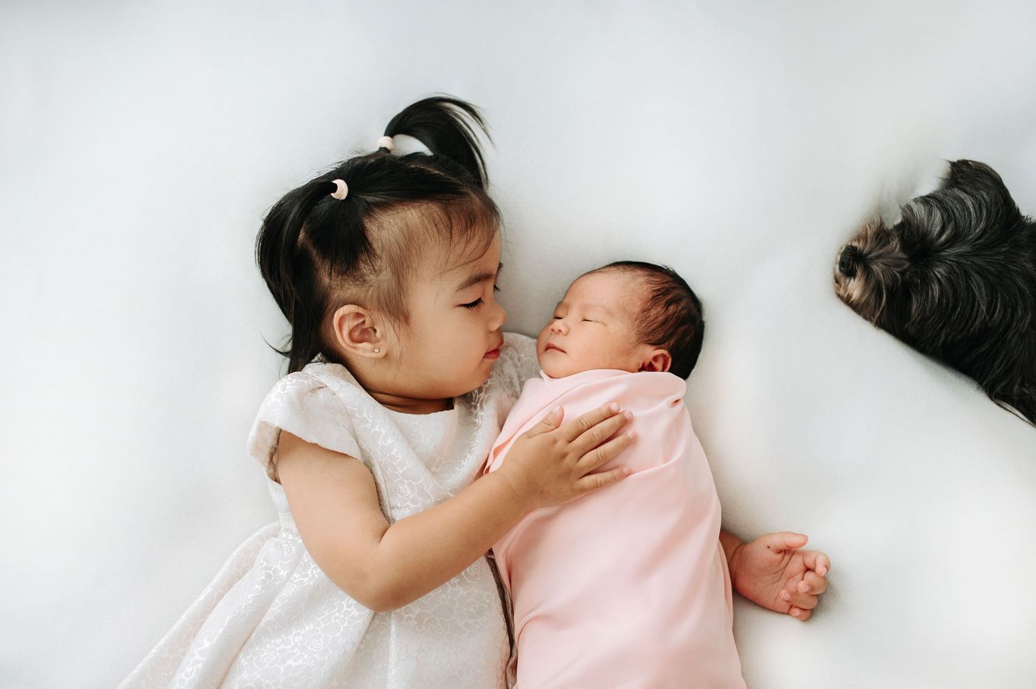 A sibling gently kisses a sleeping newborn baby while lying together on white bedding.